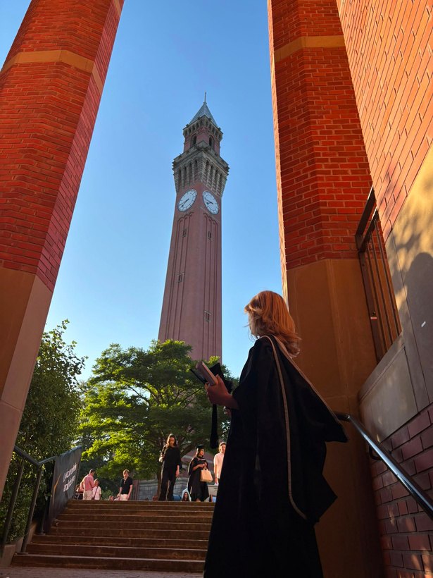 Bluebell in a graduation gown on steps below the University of Birmingham clock tower